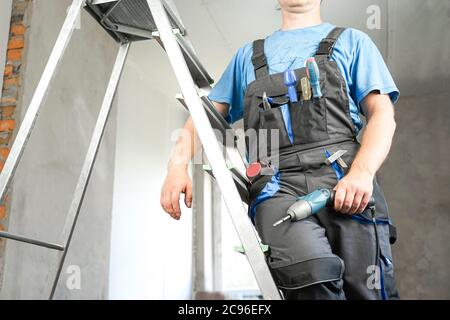 Guy in work clothes and with an electric screwdriver in his hand is standing on the stairs. Against the background of renovation in the house Stock Photo