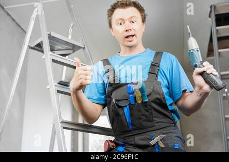 Guy in work clothes and with an electric screwdriver in his hand is standing on the stairs. Against the background of renovation in the house Stock Photo