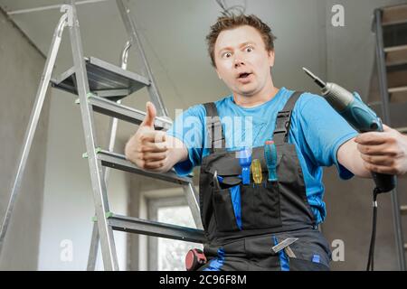 Guy in work clothes and with an electric screwdriver in his hand is standing on the stairs. Against the background of renovation in the house Stock Photo