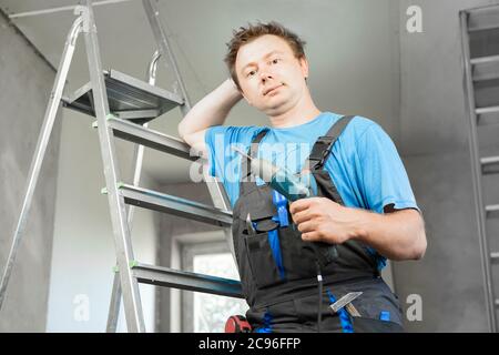 Guy in work clothes and with an electric screwdriver in his hand is standing on the stairs. Against the background of renovation in the house Stock Photo