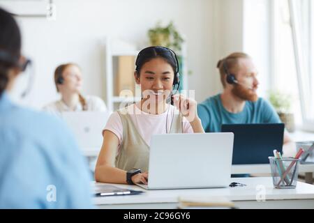 Asian young operator wearing headset sitting at workplace and using laptop she working at office with her colleagues Stock Photo