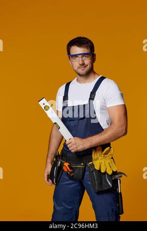 caucasian repairman worker in safety glasses and uniform holding builder level. handyman wearing tool belt isolated on orange studio background Stock Photo