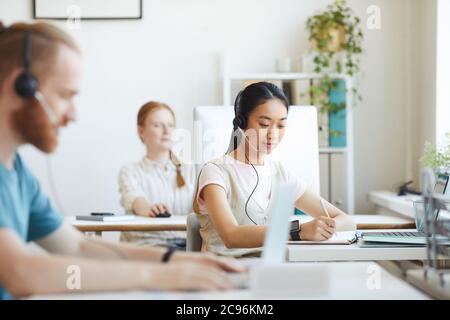 Asian young woman in headphones working at the table together with her colleagues at office Stock Photo