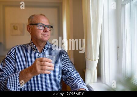 Portrait of senior businessman with glass of water looking out the window at home Stock Photo