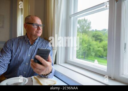 Portrait of senior businessman using phone and looking out the window at home Stock Photo