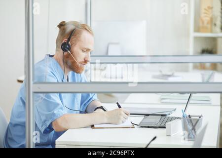 Young bearded operator in headset sitting at the table and making notes in note pad while talking on the phone Stock Photo