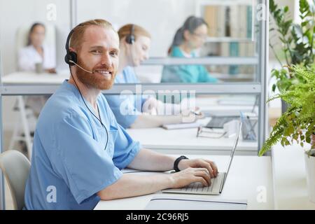 Portrait of bearded operator in headphones smiling at camera while sitting at the table and typing on laptop at office Stock Photo