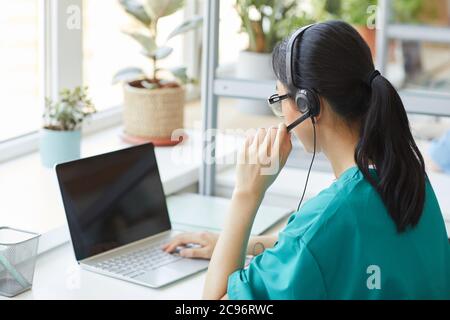Rear view of young operator in headphones talking on the phone while sitting at her workplace in front of laptop Stock Photo