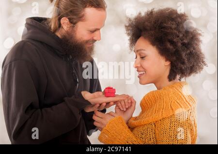 Happy man giving engagement ring in little red gift box to woman over lights background Stock Photo