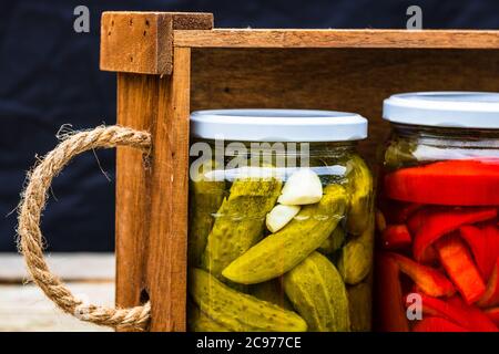 Wooden crate with glass jars with pickled red bell peppers and pickled cucumbers (pickles) isolated. Jars with variety of pickled vegetables. Preserve Stock Photo