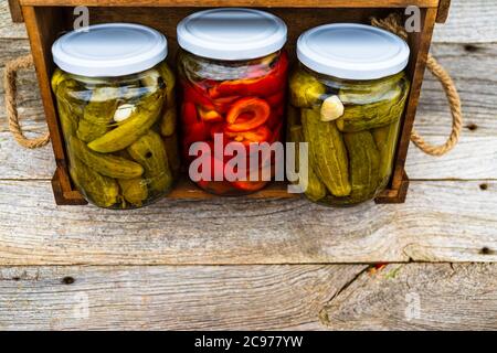 Glass jars with pickled red bell peppers and pickled cucumbers (pickles) isolated in wooden crate. Jars with variety of pickled vegetables. Preserved Stock Photo