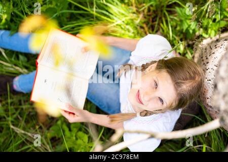 Teenage girl in white T-shirt sitting by the tree in the forest and reading interesting book. Great idea and activity to spend summer holidays. Stock Photo