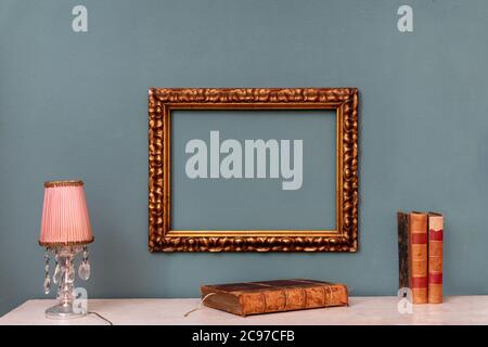 Empty gold rectangular vintage frame hanging on a green wall above a shelf with crystal drop lamp and old leather books in an interior decor concept Stock Photo
