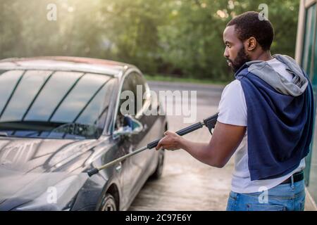 Udvikle vandfald kæmpe stor Car wash service outdoors. Side view of African guy in white t-shirt and  jeans, washing blue electric luxury car with water gun on an open air self  Stock Photo - Alamy