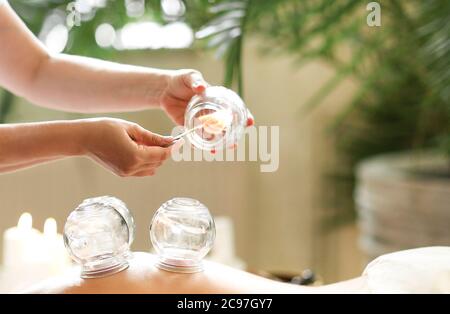 Unrecognizable person burning stick inside glass jar while performing vacuum therapy on anonymous client in spa salon Stock Photo