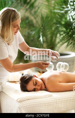 Female therapist burning stick inside glass jar while performing vacuum therapy on anonymous client in spa salon Stock Photo