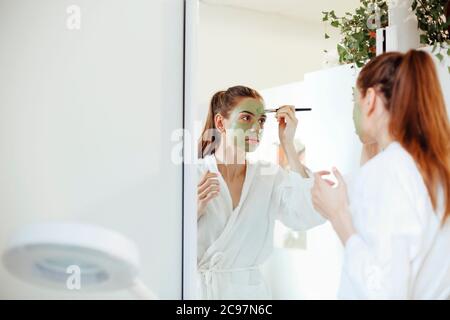 Young female in white bathrobe smearing mud mask on face while standing in front of mirror during spa procedure at home Stock Photo