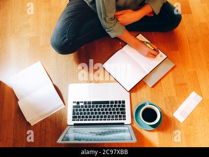 From above anonymous female freelancer sitting on floor near cup of coffee and open planner and browsing laptop while working on design project at hom Stock Photo