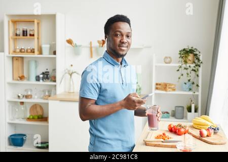 Portrait of African young man smiling at camera while drinking juice and using his mobile phone in the domestic kitchen Stock Photo