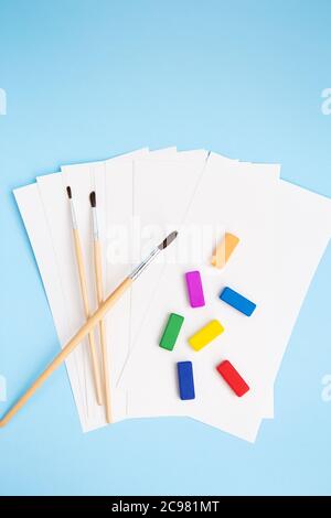 Multi-colored paints, watercolor brushes of different sizes lie together with watercolor paper on a blue background. Drawing lessons. View from above Stock Photo