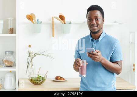 Portrait of African young guy smiling at camera and drinking fresh cocktail in the morning at home Stock Photo