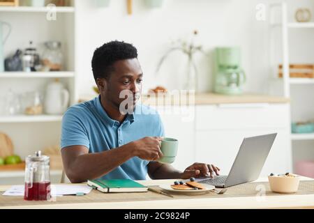 African young man sitting at the table he has breakfast and using laptop computer in the kitchen at home Stock Photo