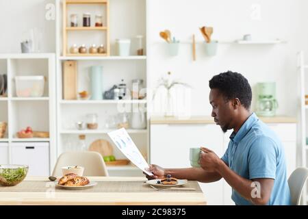 African young man sitting at the table in the kitchen he has breakfast and reading business contract Stock Photo