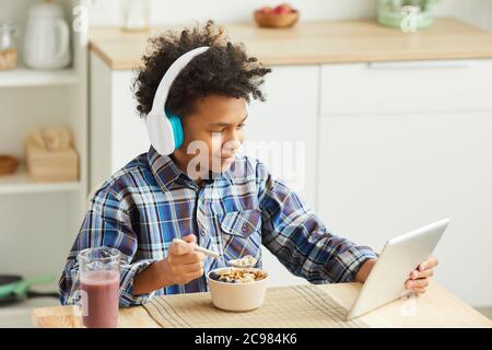 African boy with curly hair in headphones eating cereal and using digital tablet during breakfast at home Stock Photo