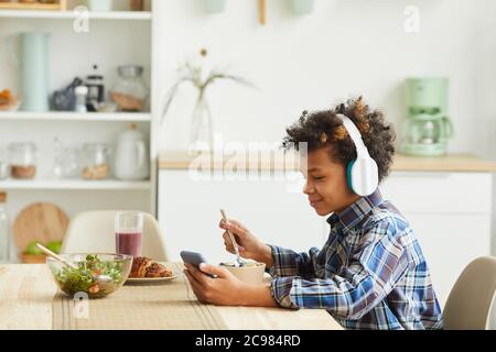 African boy in headphones using his mobile phone while eating breakfast in the kitchen Stock Photo