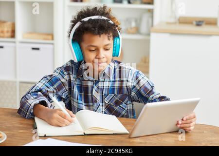 African boy in headphones using digital tablet and making notes in his notebook while sitting at the table at home Stock Photo