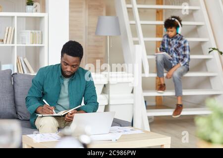 African man sitting on sofa and working online with documents and laptop with boy studying online in the background Stock Photo