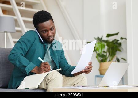 African serious businessman sitting on sofa in front of laptop and doing paperwork he working online at home Stock Photo