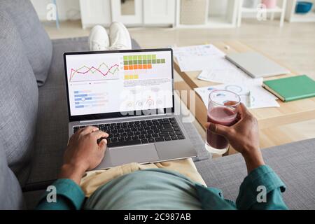 Close-up of businessman lying on sofa with fresh cocktail and examining business charts on monitor of laptop at home Stock Photo