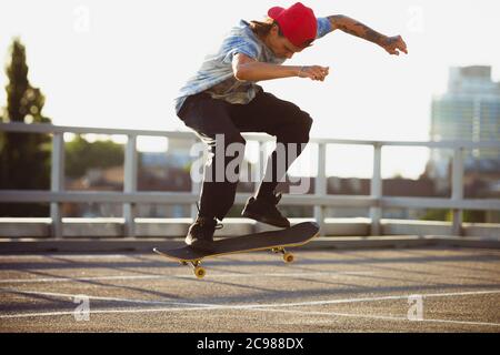 Skateboarder doing a trick at the city's street in summer's sunshine. Young man in sneakers and cap riding and skateboarding on the asphalt. Concept of leisure activity, sport, extreme, hobby and motion. Stock Photo