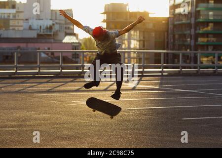 Skateboarder doing a trick at the city's street in summer's sunshine. Young man in sneakers and cap riding and skateboarding on the asphalt. Concept of leisure activity, sport, extreme, hobby and motion. Stock Photo