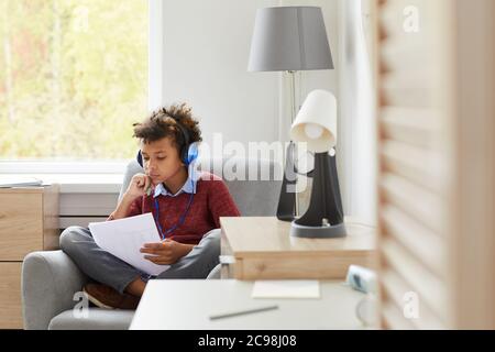 African boy in headphones sitting on armchair with papers he learning lessons or preparing for the exam at home Stock Photo