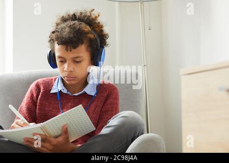 Serious African boy in headphones sitting on armchair and making notes in textbook he preparing for the lessons at home Stock Photo