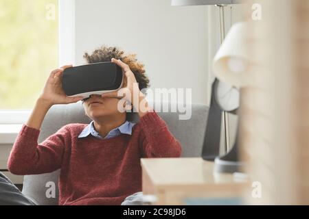 African boy wearing virtual glasses sitting on armchair and playing in virtual games at home Stock Photo