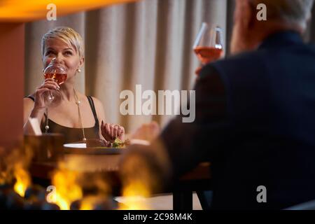 Happy gray-haired elderly lady in black dress toasting with glass of champagne to male in elegant restaurant, look at him. Rear view on man in tuxedo.