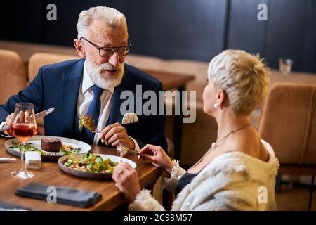 Pretty lady and elderly grey haired man in tuxedo having friendly conversation in restaurant. Colleagues after work discuss and eat Stock Photo