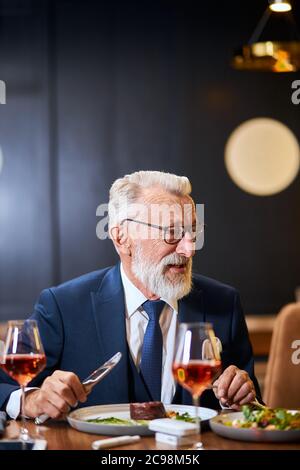 Bearded elderly businessman in glasses on a meeting in a restaurant, having a conversation with business partner and taking a meal. Man eats from beau Stock Photo