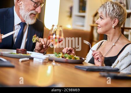 Elderly male and female chatting, looking at each other. Bearded man in tuxedo and glasses talking, woman looks at him. Horizontal view. Stock Photo