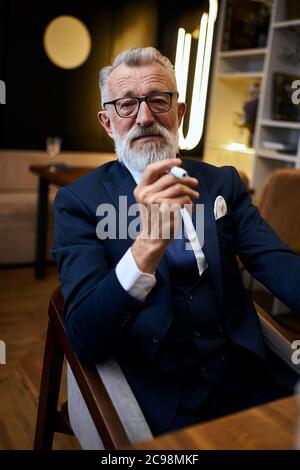 Handsome grey-haired senior man in glasses in suit smoke IQOS in restaurant, explore cigarette holding in hands Stock Photo