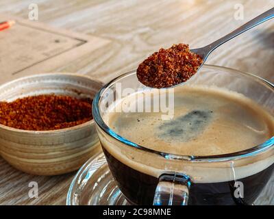Spoon full of brown sugar over a cup of black morning coffee in a glass cup, on a light brown wood table, start for coffee Stock Photo