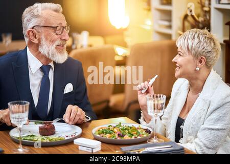 Elderly couple have dinner in rich restaurant while using IQOS, e-cigarette. Handsome man in tuxedo smile, look at woman and speak. Stock Photo