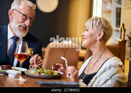 Elderly elegant people taking meal in beautiful restaurant. Free and modern, using iqos, e-cigarettes. Caucasian grey-haired and beared male in tuxedo Stock Photo
