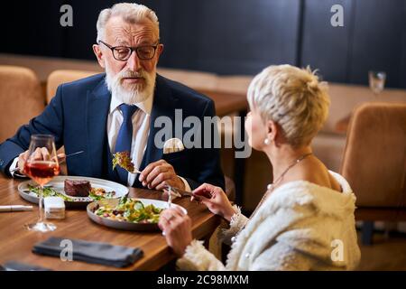 Pretty lady and elderly grey haired man in tuxedo having friendly conversation in restaurant. Colleagues after work discuss and eat Stock Photo