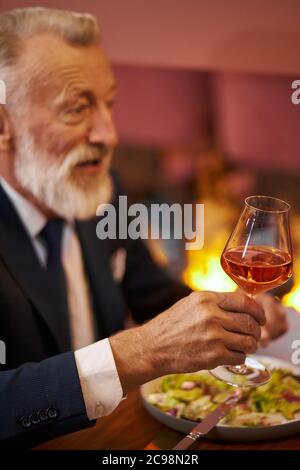 Senior elegant bearded and gray-haired man in tuxedo with glass of champagne sit in restaurant and laugh, look at person opposite. Background fireplac Stock Photo