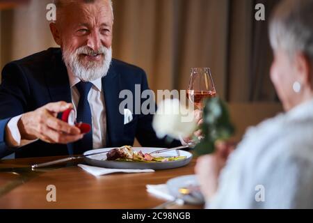 Senior man happily looks at woman waiting answer for marriage proposing. Elegant male in suit smile holding a ring, woman hold white rose. Love, relat