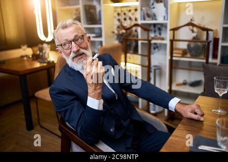 Attractive older man enjoys smoking electronic cigarette in restaurant. Senior man using e-cigarette, IQOS Stock Photo
